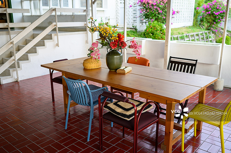 一家舒适的半户外露台餐厅照片，设有木桌、混搭椅子和花卉中心装饰，周围环绕着郁郁葱葱的热带绿植图片素材