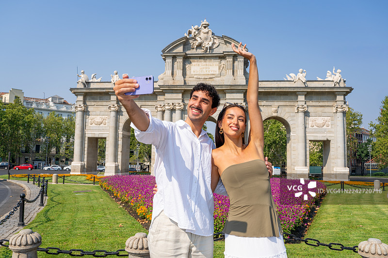 年轻的多元文化情侣在西班牙马德里阿尔卡拉门前，开心地用智能手机自拍，庆祝他们的暑假并享受旅游的乐趣。图片素材