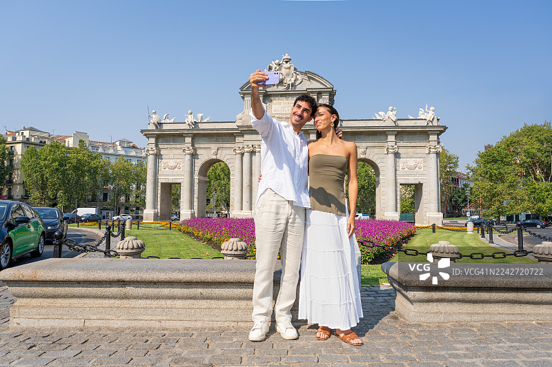 一对快乐的游客情侣在马德里历史悠久的阿尔卡拉门前自拍，享受阳光明媚的城市度假、观光和夏日假期。西班牙马德里。图片素材