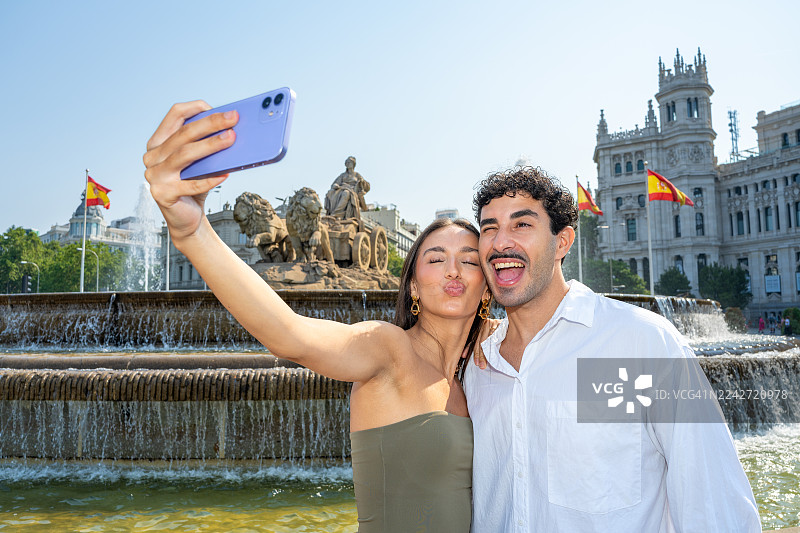一对年轻的游客网红情侣正在度假,他们对着手机自拍,做出搞怪的表情,男方眨着眼睛,女方撅着嘴,背景是西班牙马德里标志性的西贝莱斯喷泉和西贝莱斯宫。图片素材