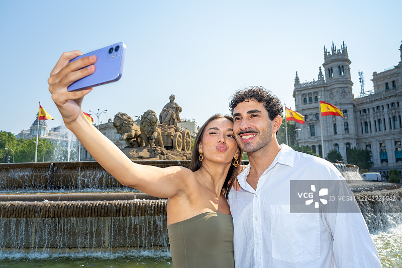 一对年轻情侣在马德里西贝莱斯喷泉前开心自拍,她俏皮地眨眼嘟嘴,在标志性喷泉和历史悠久的城市地标前一同享受阳光明媚的夏日假期。西班牙马德里图片素材