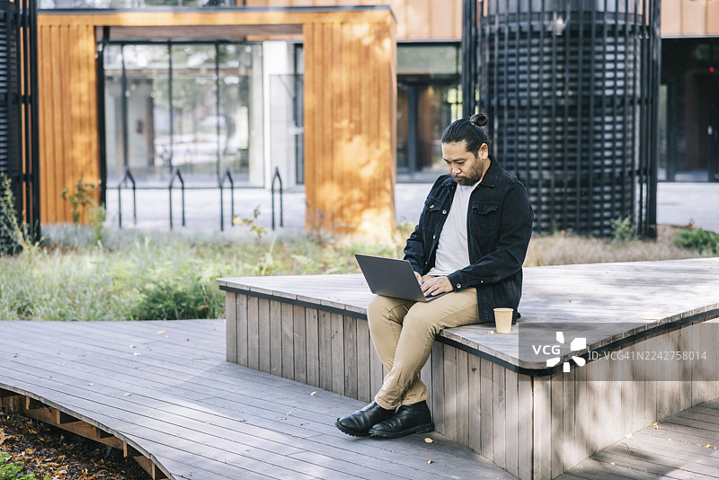 一名男子在城市公园的户外使用笔记本电脑工作，他坐在一张木长椅上，旁边放着一杯咖啡图片素材