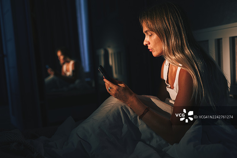 夜间，在安静昏暗的卧室灯光下，一名女子在床上使用智能手机图片素材