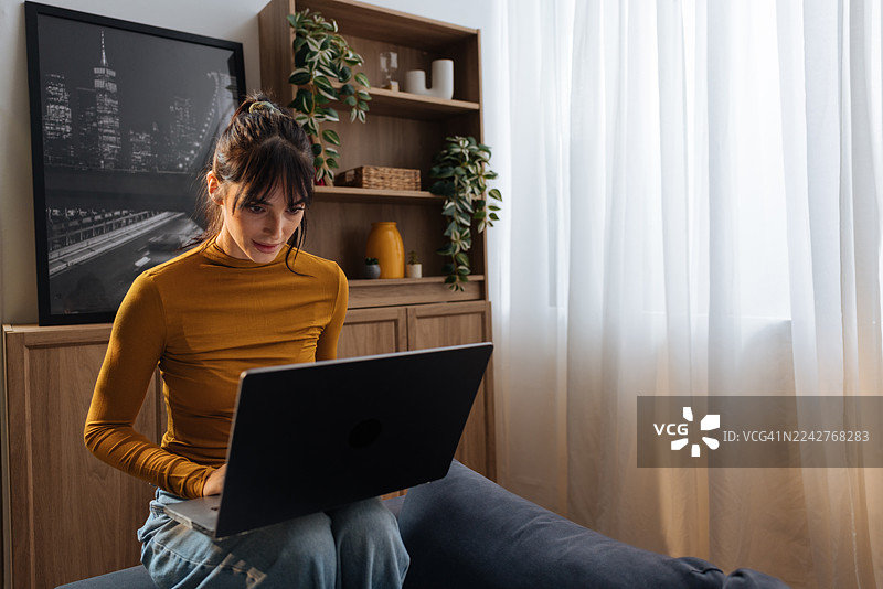 女人在家中舒适的客厅里，坐在沙发上使用笔记本电脑，周围有植物和书架图片素材