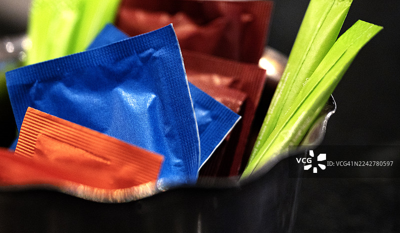 特写镜头：碗里装着各种颜色（蓝、绿、红）的糖包和甜味剂包装。图片素材