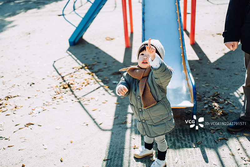 一个可爱的亚洲小女孩，在晴朗的冬日里，在户外的游乐场玩耍，她抬头指向天空，和她的父亲在公园里玩得很开心。家庭生活方式。父女时光。户外乐趣。图片素材