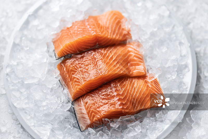 三片新鲜三文鱼，摆放在盛满碎冰的白盘上。三文鱼鲜亮的橙色与冰冷背景形成鲜明对比，一小枝迷迭香点缀其间，暗示着烹饪的准备。图片素材