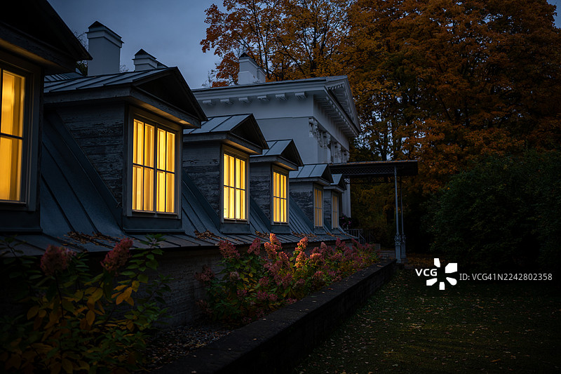 秋日阴云笼罩的傍晚，户外花园里的舒适灌木丛旁，房屋窗户透出温馨的灯光。图片素材