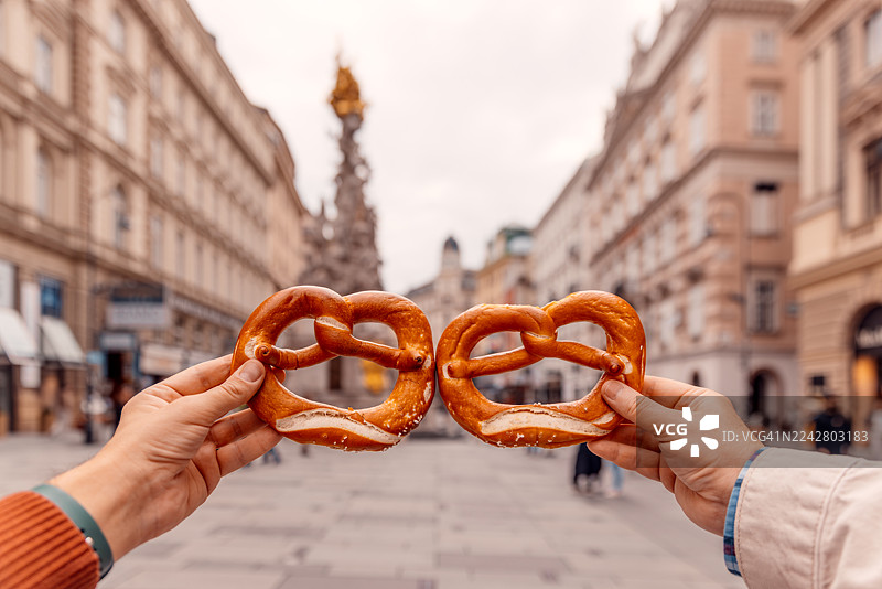 从个人视角看，在奥地利维也纳历史悠久的老城区格拉本购物街上，两个人正拿着椒盐卷饼。图片素材