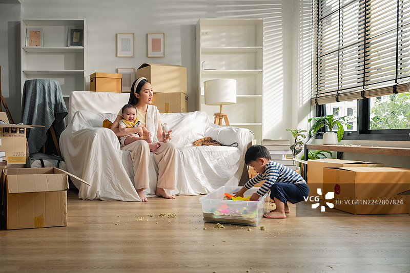 年轻的亚洲母亲和她的两个孩子（男孩和女婴）在搬家过程中，兴高采烈地拆着纸箱并摆放家具，在新家搬迁的喜悦中享受着轻松的玩耍时刻，营造出宁静的生活方式。图片素材
