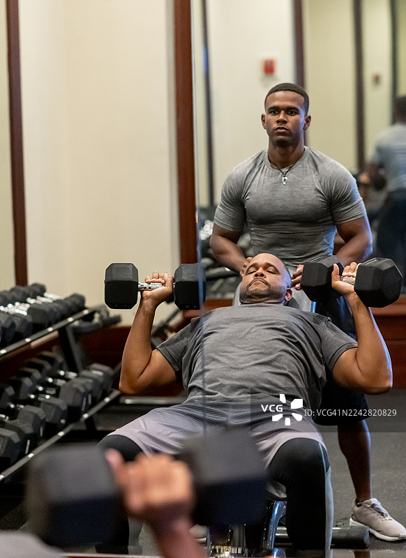 两位非裔美国男性，一位是父亲，一位是成年儿子，正在健身房一起锻炼，保持健康的生活方式，体现家庭团聚图片素材