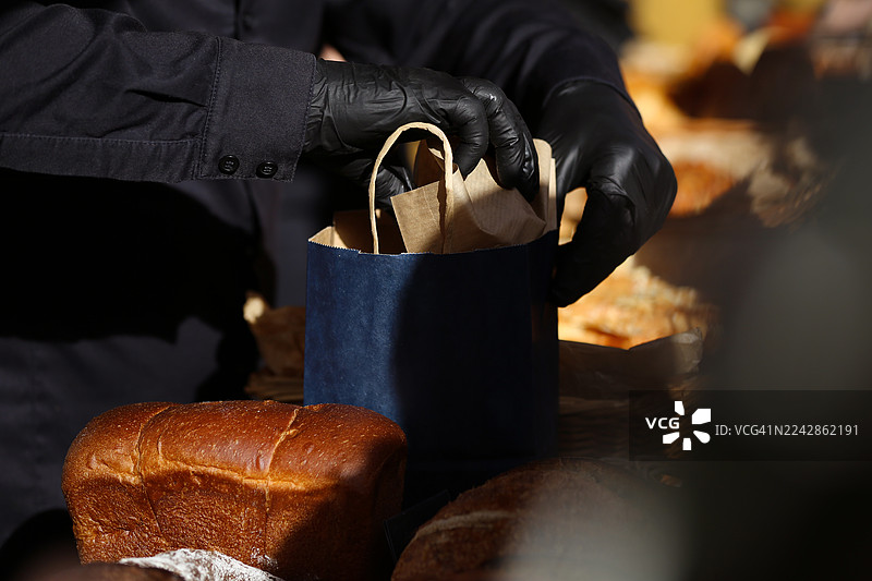 在路边咖啡馆，烘焙师的手将新鲜的烤面包棒装进棕色纸午餐袋的特写图片素材