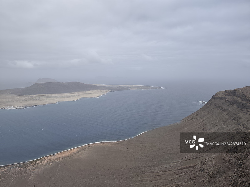 从空中俯瞰，在多云的天空下，崎岖的海岸线与平静的大海构成了一幅荒凉而美丽的岛屿风景画，地点是西班牙拉斯帕尔马斯。图片素材