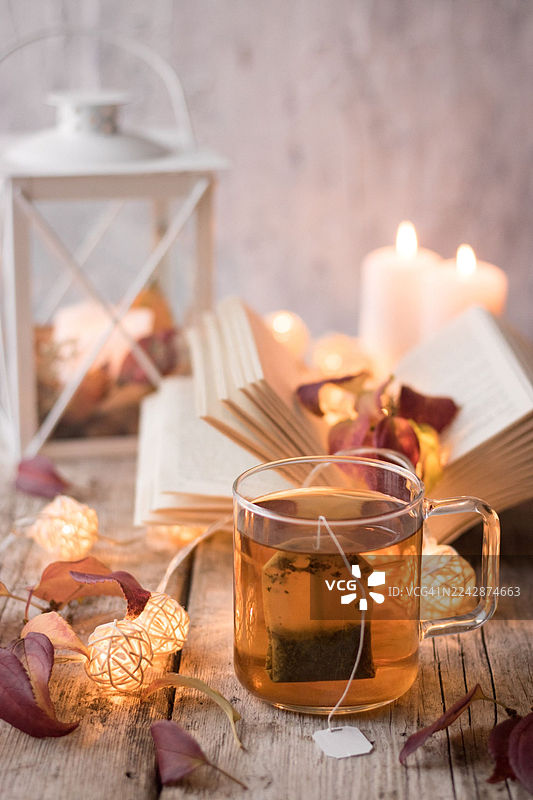 舒适的秋日景象，原木桌上摆放着冒着热气的茶杯、打开的书籍、蜡烛和装饰灯，营造出温暖放松的氛围图片素材