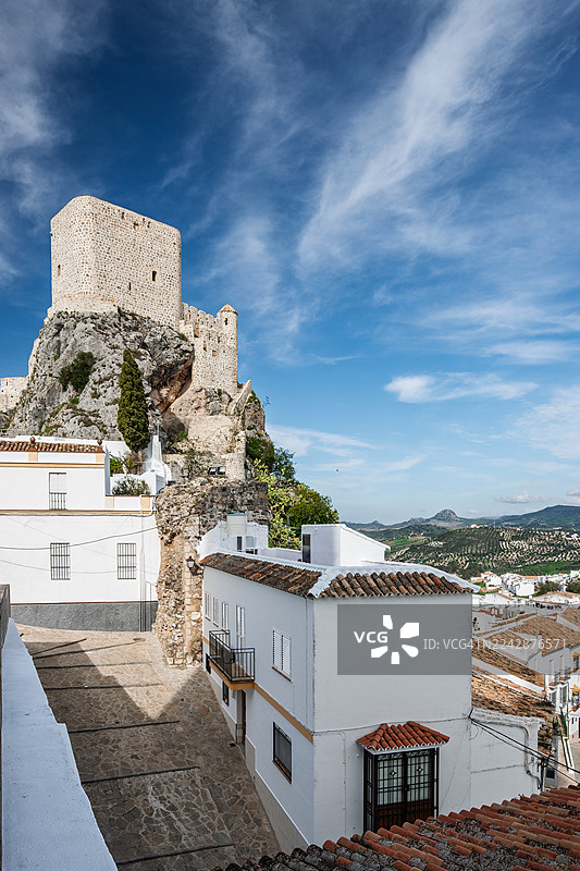 西班牙安达卢西亚，一座历史悠久的城堡矗立在岩石峭壁上，俯瞰着传统白色村庄，背景是戏剧性的蓝色天空和缕缕白云。图片素材