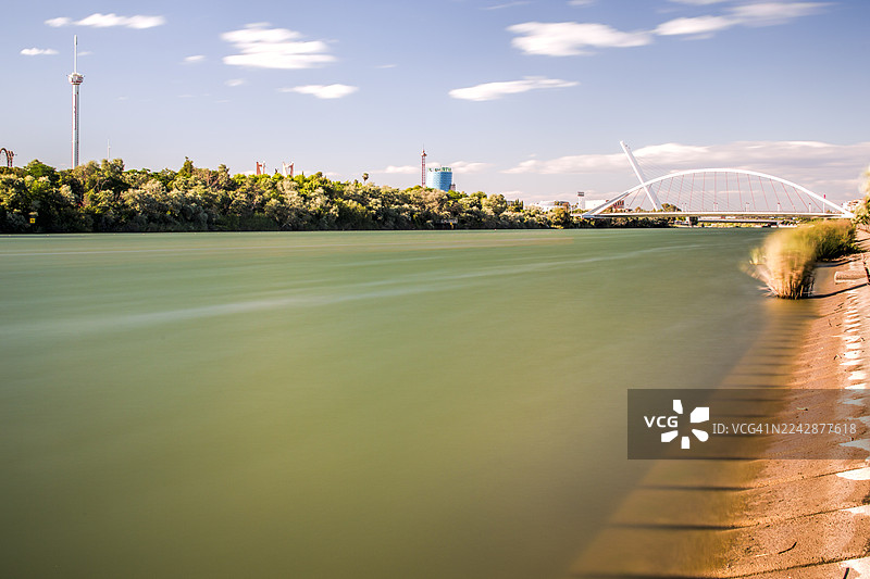 西班牙塞维利亚，长曝光拍摄的宁静河流、现代建筑和茂密树木在蓝色天空下的风景图片素材