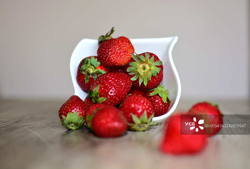 特写镜头：新鲜成熟的草莓从白色碗中溢出，散落在木桌上，鲜艳的红色和天然纹理清晰可见图片素材
