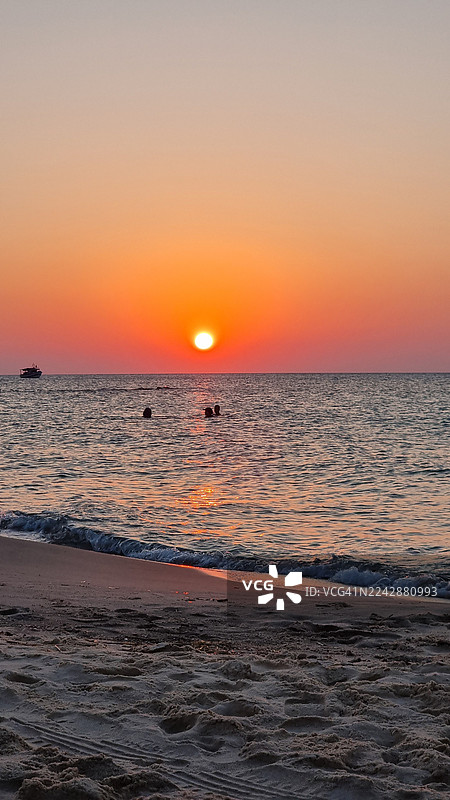 日落时分，海景与生动的晚霞交相辉映，海中有游泳的人，远处还有一艘船。图片素材