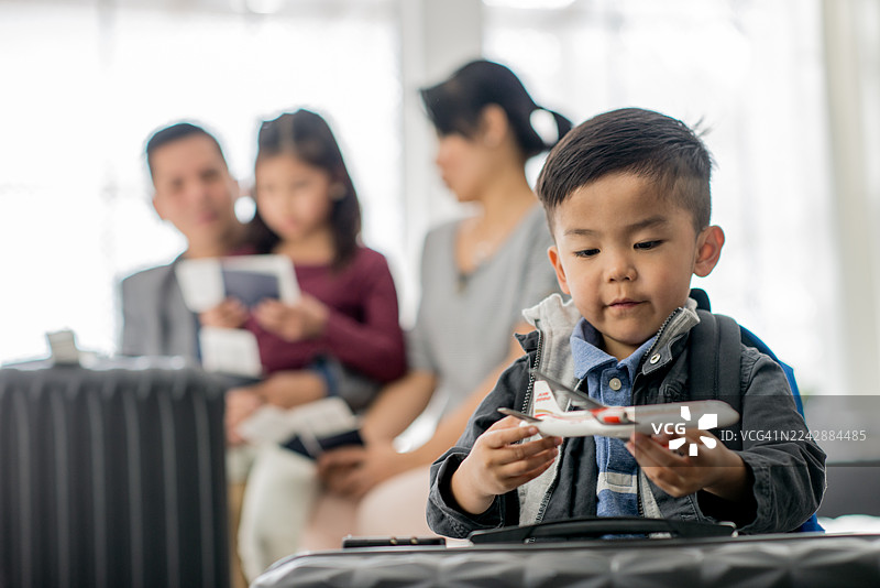 一个亚裔小男孩在为旅行做准备的环境中玩玩具飞机，家人在一旁观看图片素材