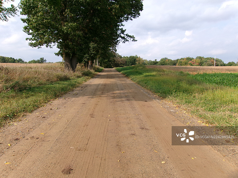一条空旷的土路蜿蜒穿过田野和树林，天空布满云彩，地点为美国俄克拉荷马州巴尔奥克。图片素材