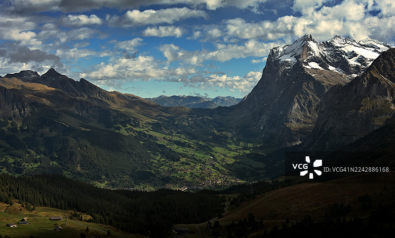 雄伟的阿尔卑斯山风光，有白雪皑皑的山峰、翠绿的山谷和坐落于戏剧性天空下的村庄，瑞士格林德瓦尔德图片素材