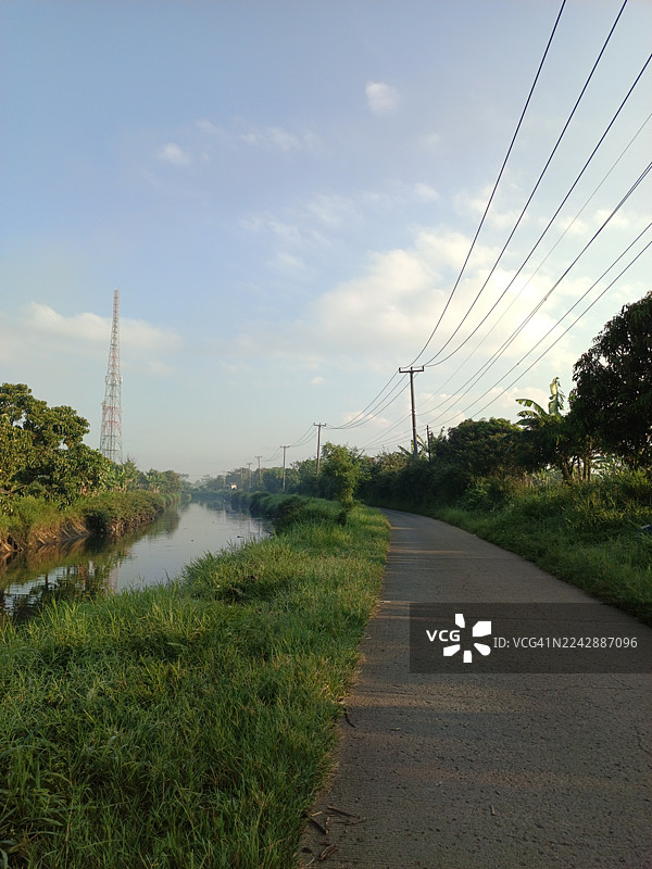 绿树成荫，运河穿行，天空晴朗，远处有通讯塔和电线，一条空旷的道路图片素材