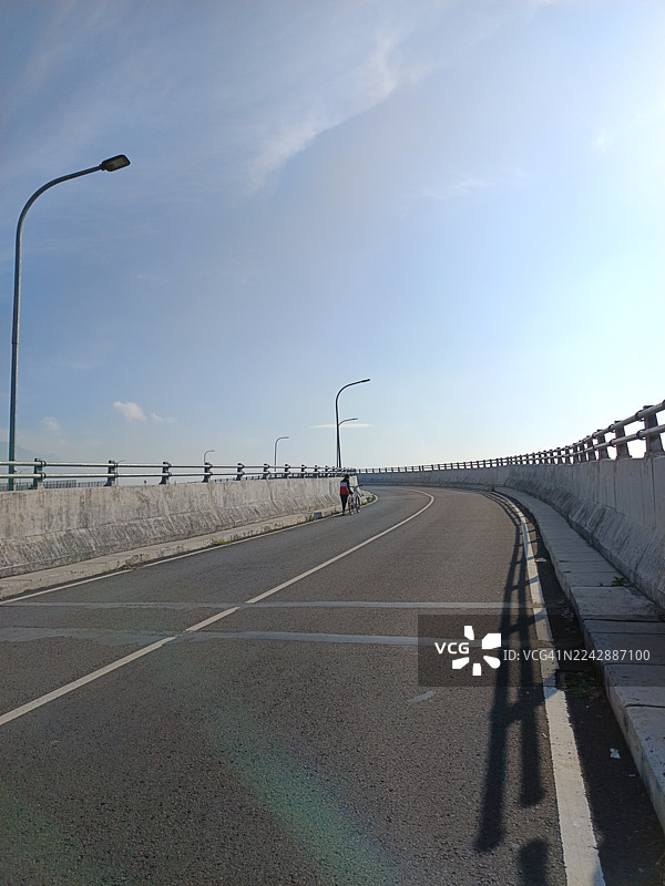 晴朗蓝天下，一个人在印尼万隆的立交桥上推着自行车行走，下方是空旷的沥青路。图片素材