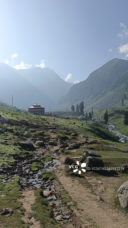 巴基斯坦库姆拉特山谷，郁郁葱葱的绿色山谷风景，一条蜿蜒的土路，一座传统建筑，在明媚的天空下。图片素材