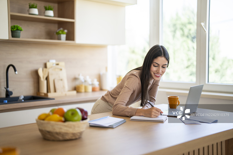 年轻女子在现代厨房里用笔记本电脑记笔记，在家办公图片素材