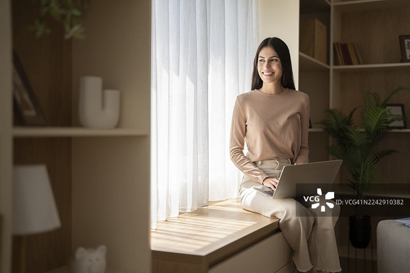 年轻女子在家靠窗用笔记本电脑工作图片素材