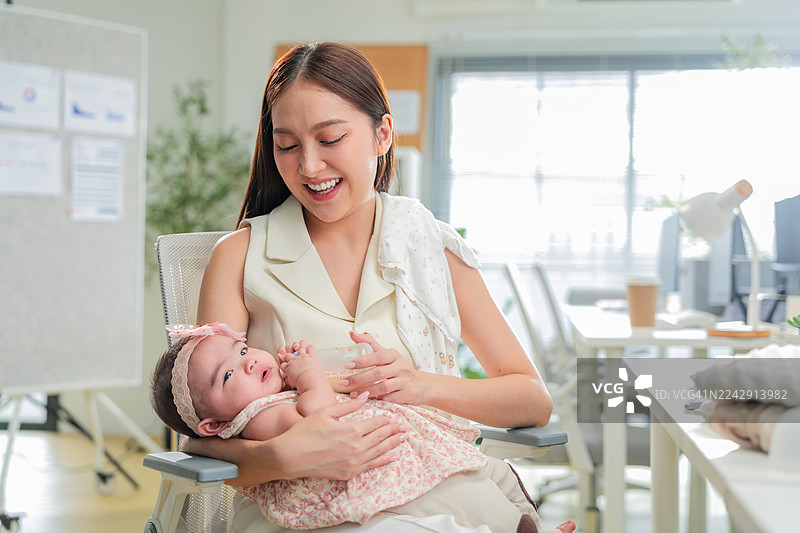 年轻、现代生活方式的亚洲职业女性在办公室高效地处理多项任务，同时充满爱意地喂养女儿婴儿喝奶，展现了职业奉献精神和母性关怀图片素材