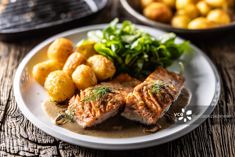 浅盘中盛放着两片烤三文鱼，淋上蘑菇酱，撒上莳萝，旁边配有烤土豆和新鲜的生菜沙拉。图片素材