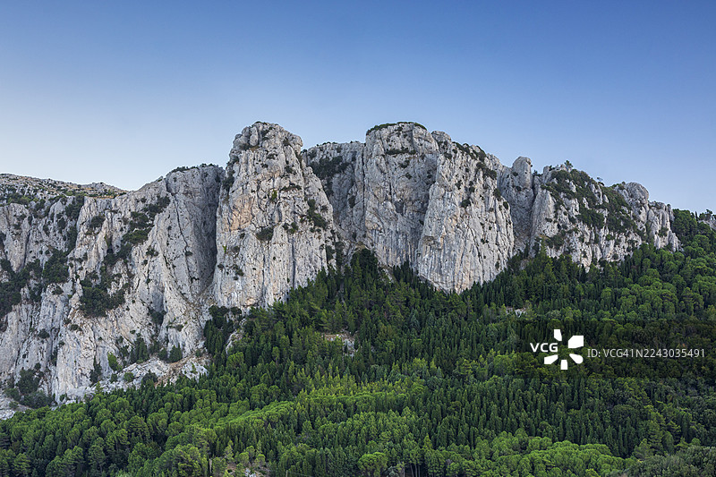 意大利卡诺洛，雄伟的粗糙岩层在晴朗的蓝天下，底部有郁郁葱葱的绿色森林，低角度仰视图片素材