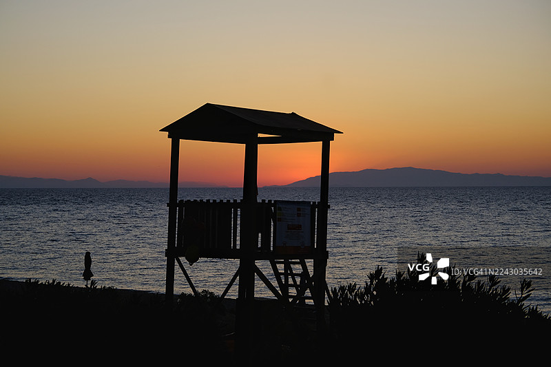 希腊斯卡拉，海边救生员小屋在生机勃勃的日落天空下映衬出剪影，远方的山脉在地平线上若隐若现。图片素材