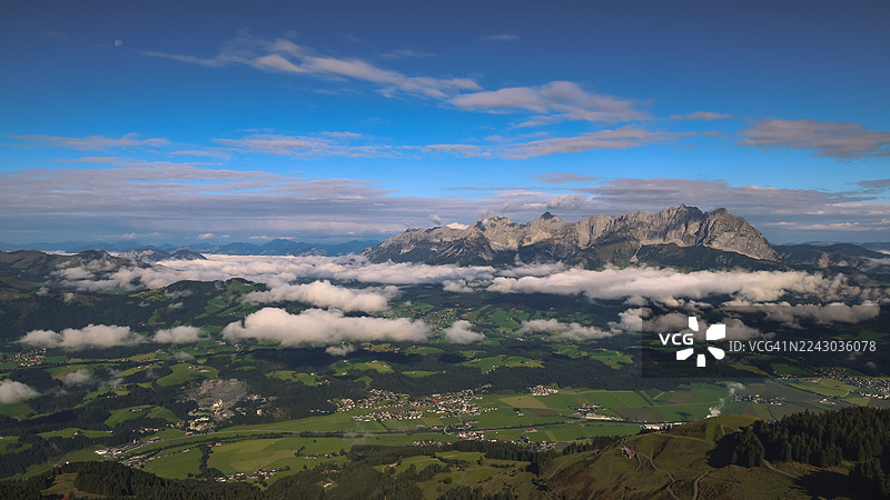 奥地利，蓝色天空点缀着零星云朵，令人惊叹的航拍景象，群山环绕的山谷中云雾缭绕，点缀着小村庄图片素材