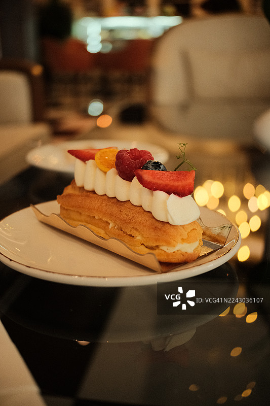 白色盘子上的美味水果闪电泡芙特写，在背景虚化灯光的餐厅环境中，展示着鲜艳的浆果和奶油图片素材