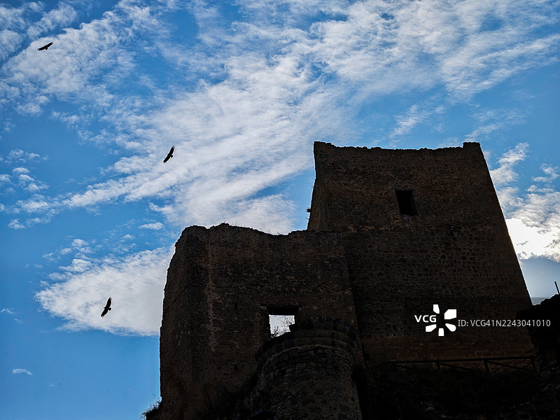 从低角度看，鸟儿飞过古老的堡垒，背景是戏剧性的蓝天白云，西班牙图片素材