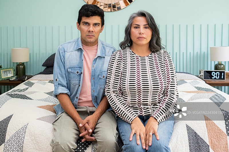 在墨西哥城，一对拉丁裔夫妇在争吵后，严肃地看着镜头，坐在他们的床上。图片素材