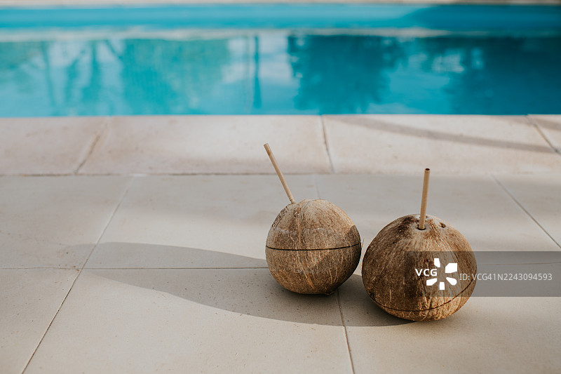 两个椰子插着吸管，放在热带气候的游泳池旁边。图片素材