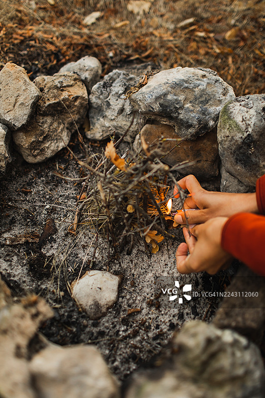秋季户外，一人在石头营火堆旁收集细小的树枝并点燃篝火图片素材