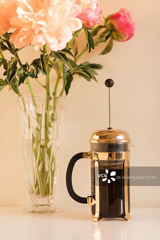 金色与黑色的法式压滤壶，旁边放着一杯黑咖啡、一个深绿色马克杯和一束粉色渐变牡丹花，花插在水晶花瓶中，置于晨光下。图片素材