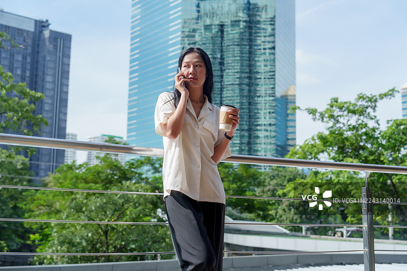 年轻的亚洲专业人士在户外，一位女商人在城市阳台上，背景是城市天际线，手里拿着咖啡和智能手机，在明亮的日光下，情绪自信，风格休闲。图片素材