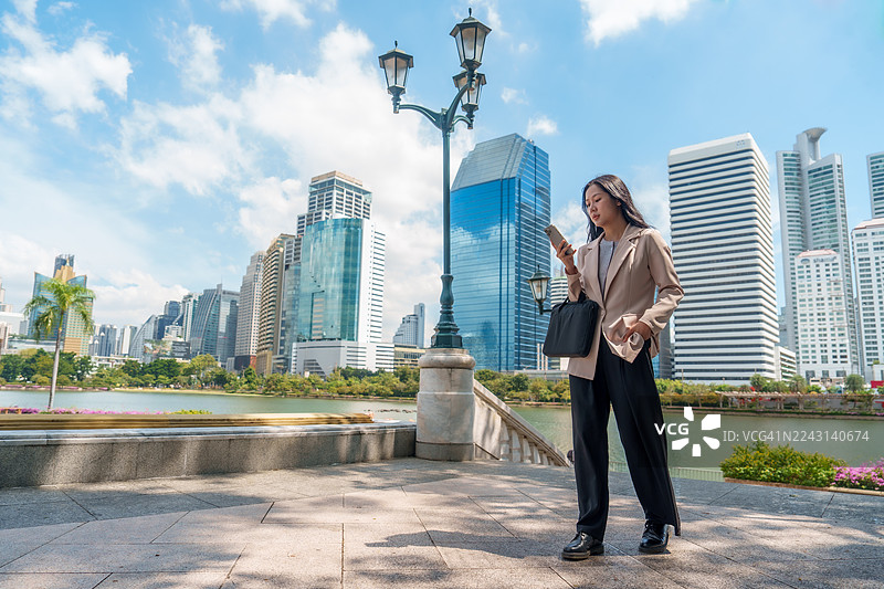 一位优雅的女商人在阳光明媚的白天，在城市公园里，身穿休闲西装外套，手拿皮包，身旁有智能手机，眺望着现代建筑的滨水摩天大楼天际线。她心情自信，氛围宁静祥和。图片素材