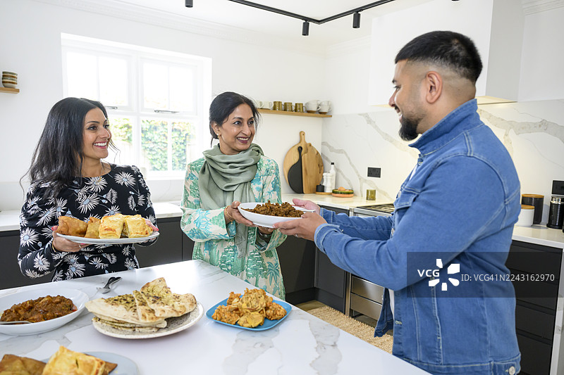英裔巴基斯坦家庭在厨房准备餐食，一名男子从一位年长女性手中接过盘子图片素材