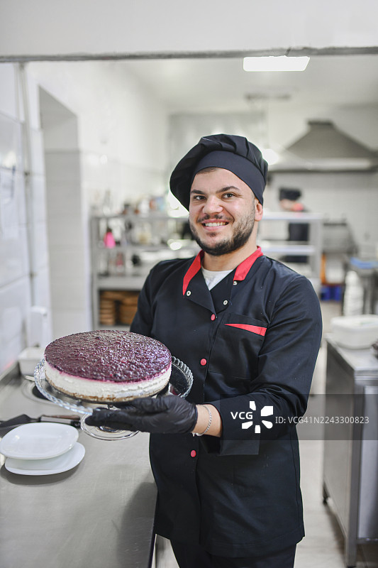 身穿黑色制服的厨师面带微笑，在现代厨房工作室展示着装饰有浆果的蛋糕图片素材