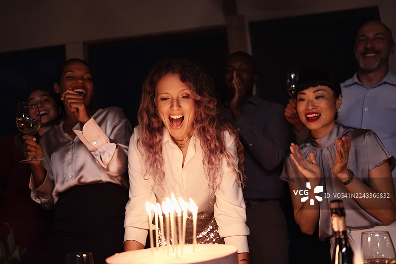 朋友们在夜晚派对上，伴随着欢声笑语和碰杯声，庆祝生日，蛋糕上插着蜡烛。图片素材