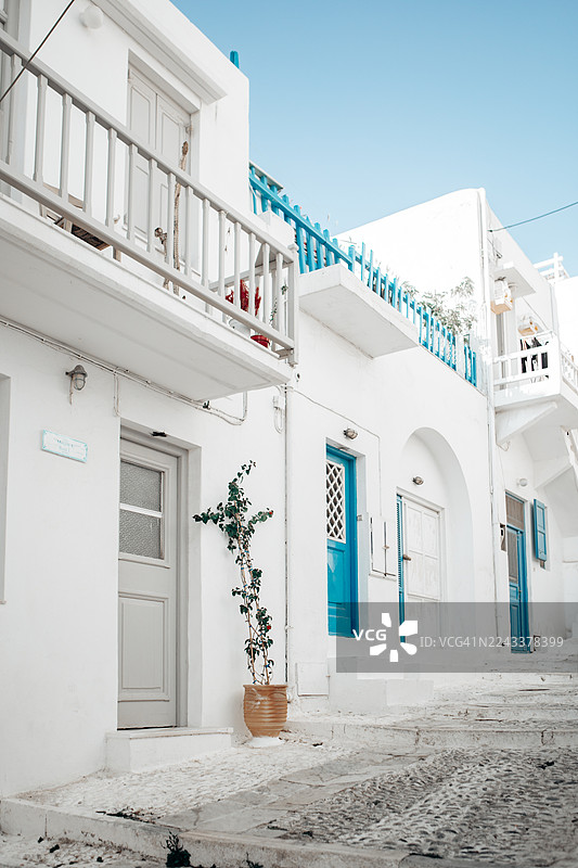 这张图片展示了希腊米科诺斯岛的一条街道，该岛以其传统的基克拉迪建筑而闻名，房屋是白色的，并配有蓝色的门和栏杆等鲜艳的细节。图片素材
