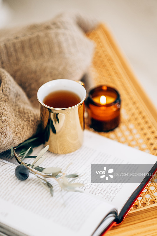 舒适纹理毯子和编织凳子上，放着一个金色的反光马克杯，里面装着茶，旁边还有一本打开的书、一支点燃的蜡烛和一根橄榄枝。图片素材