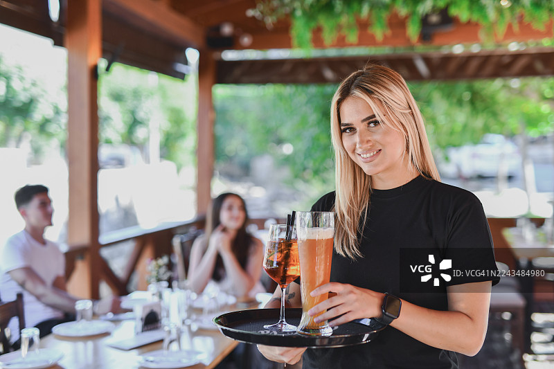 面带微笑的女服务员在餐厅露台上为顾客送上饮品，背景中是她的朋友们图片素材
