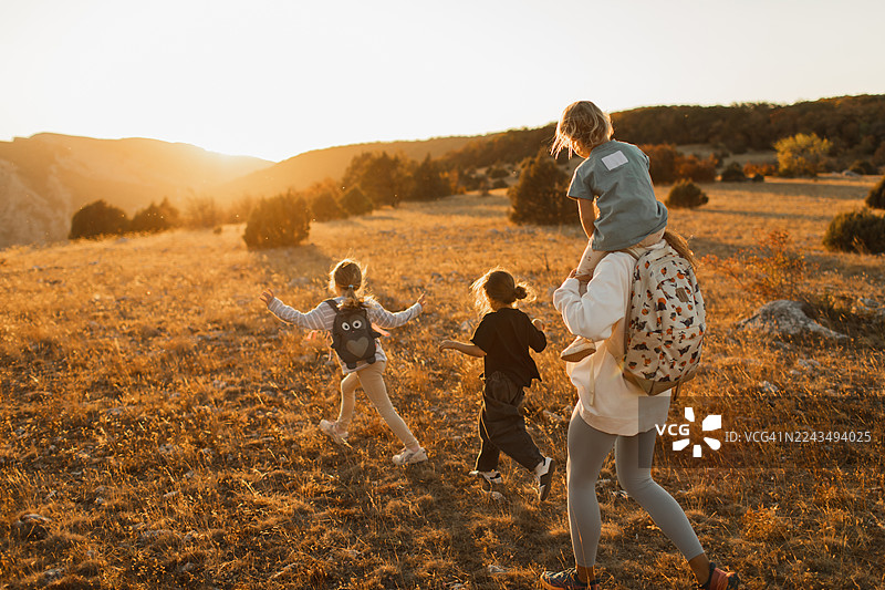 夕阳下的家庭户外探险：孩子们在前面奔跑，妈妈则将一个孩子扛在肩上图片素材